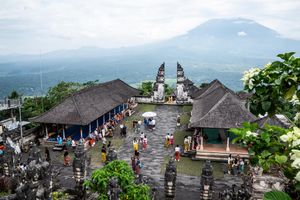Aerial view of a traditional Balinese temple complex with ornate gates, visitors in colorful attire, and a misty volcanic mountain backdrop.