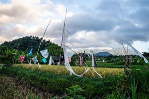 Colorful flags and streamers on tall poles in lush green rice fields with mountains and cloudy sky backdrop.