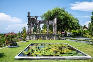 Ancient stone arch ruins reflected in a lily pond garden under a bright blue sky in Bali.