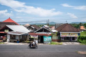 Motorcyclist rides past traditional Balinese buildings on a quiet street with lush green hills and blue sky in background.