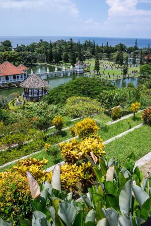 Aerial view of Taman Ujung water palace gardens with yellow flowers, pavilions, and ocean backdrop in Bali.