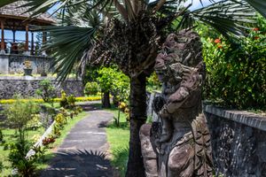 Ornate Balinese stone statue beside a palm tree in a lush tropical garden with a stone pathway.