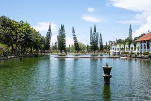 Serene garden pond with tall cypress trees, lush greenery, and a white colonial building under a bright blue sky.