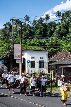 Traditional Balinese procession with white-clad musicians marching down a tropical road with lush green hills.