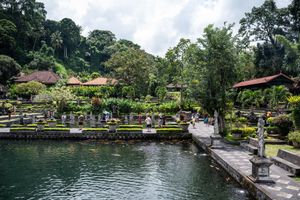 Lush Balinese water temple garden with serene pond, traditional pavilions, tropical trees, and stone statues under cloudy skies.