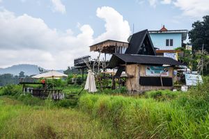 Rustic wooden stilt house with black A-frame roof surrounded by lush green fields and mountains in rural Southeast Asia.
