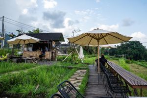 Outdoor cafe with beige umbrellas, wooden deck, lush greenery, and rustic seating area under a partly cloudy sky.