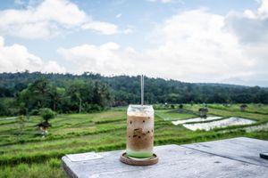 Iced coffee drink on a wooden table overlooking lush green rice terraces and cloudy sky in Bali.