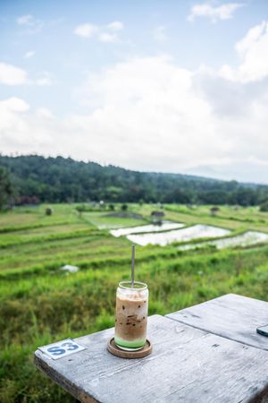 Iced coffee drink on wooden table overlooking lush green rice fields under blue sky in Bali.