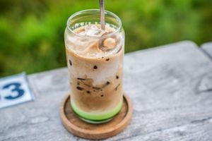Close-up of iced coffee with milk on wooden coaster, served with metal straw outdoors.
