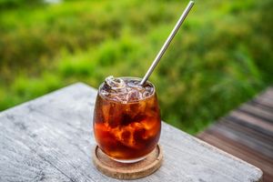 Iced coffee cocktail in a glass with metal straw on wooden coaster, set against lush green background.