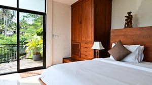 Bright tropical villa bedroom with wooden furniture, white bedding, and a balcony overlooking greenery.