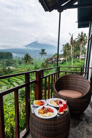 Breakfast table on a balcony overlooking lush Bali rice terraces and misty volcanic mountain backdrop.