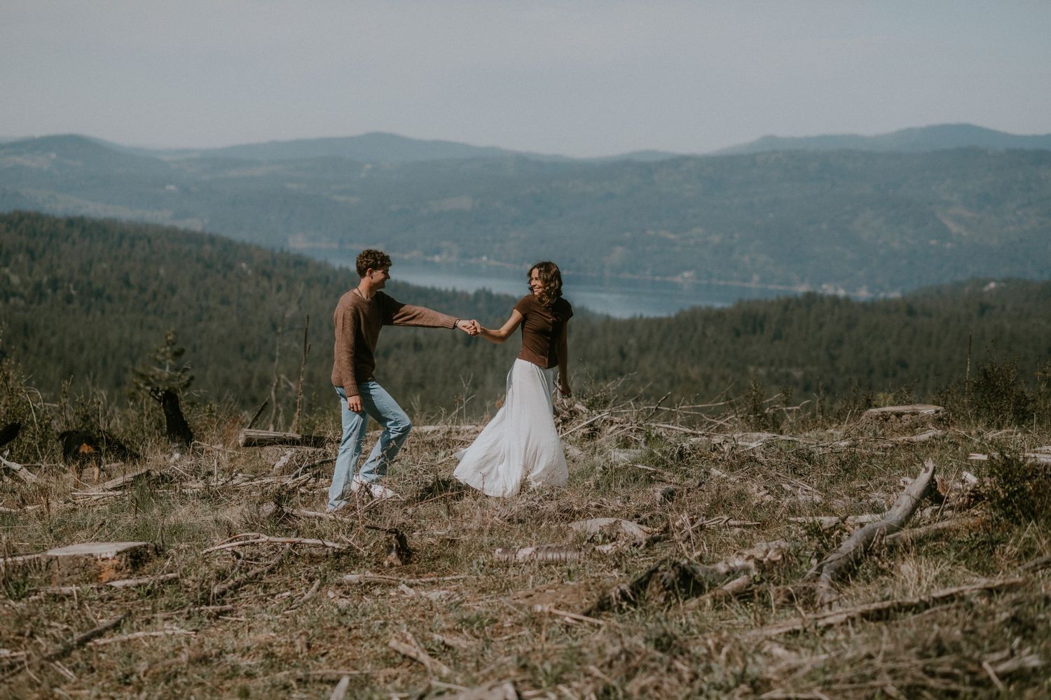 Couple holding hands dancing on a scenic mountain hilltop with forest and lake views in wedding attire.