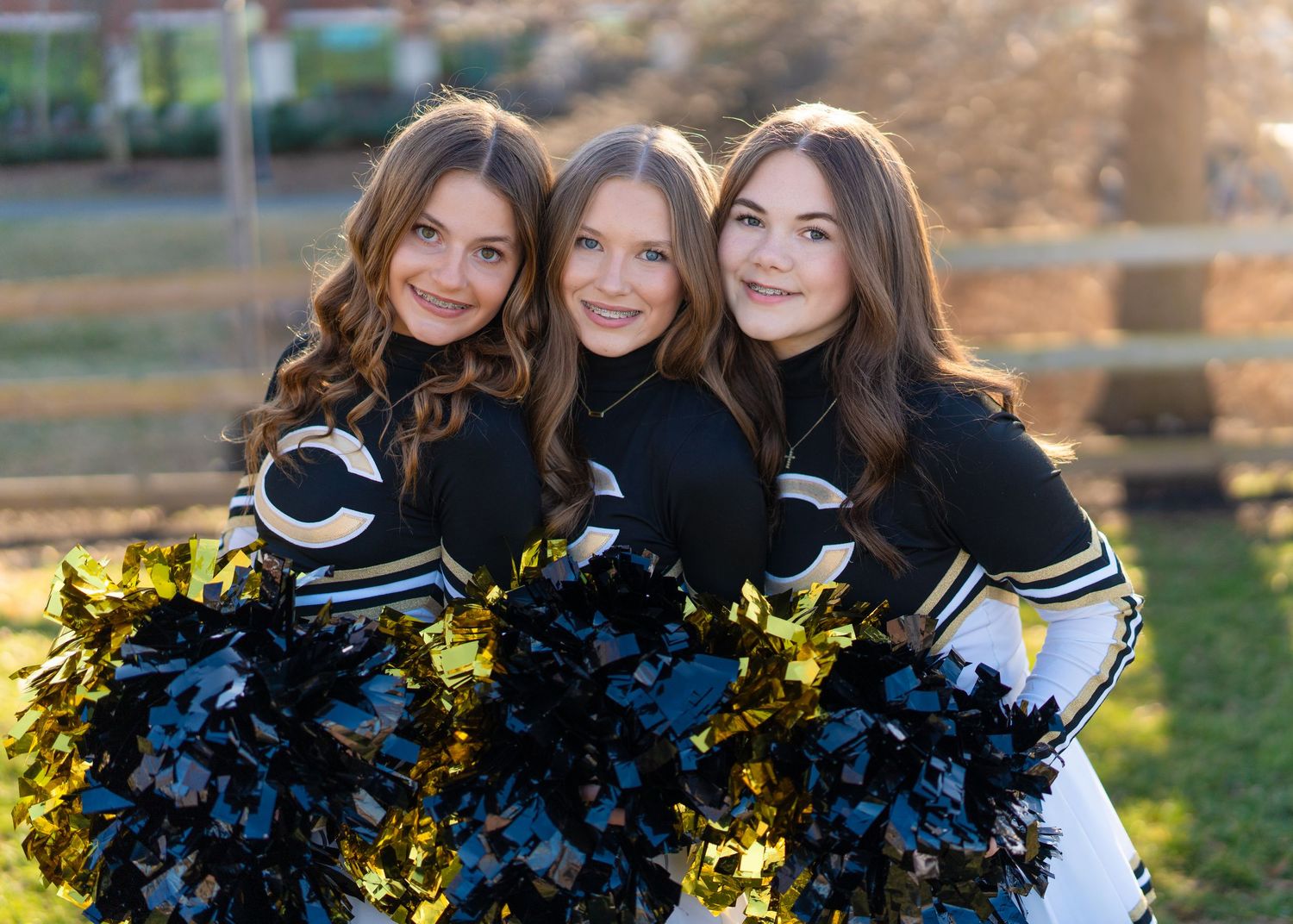 Three Christiana Middle School cheerleaders in black and gold uniforms pose together holding pom-poms outdoors.