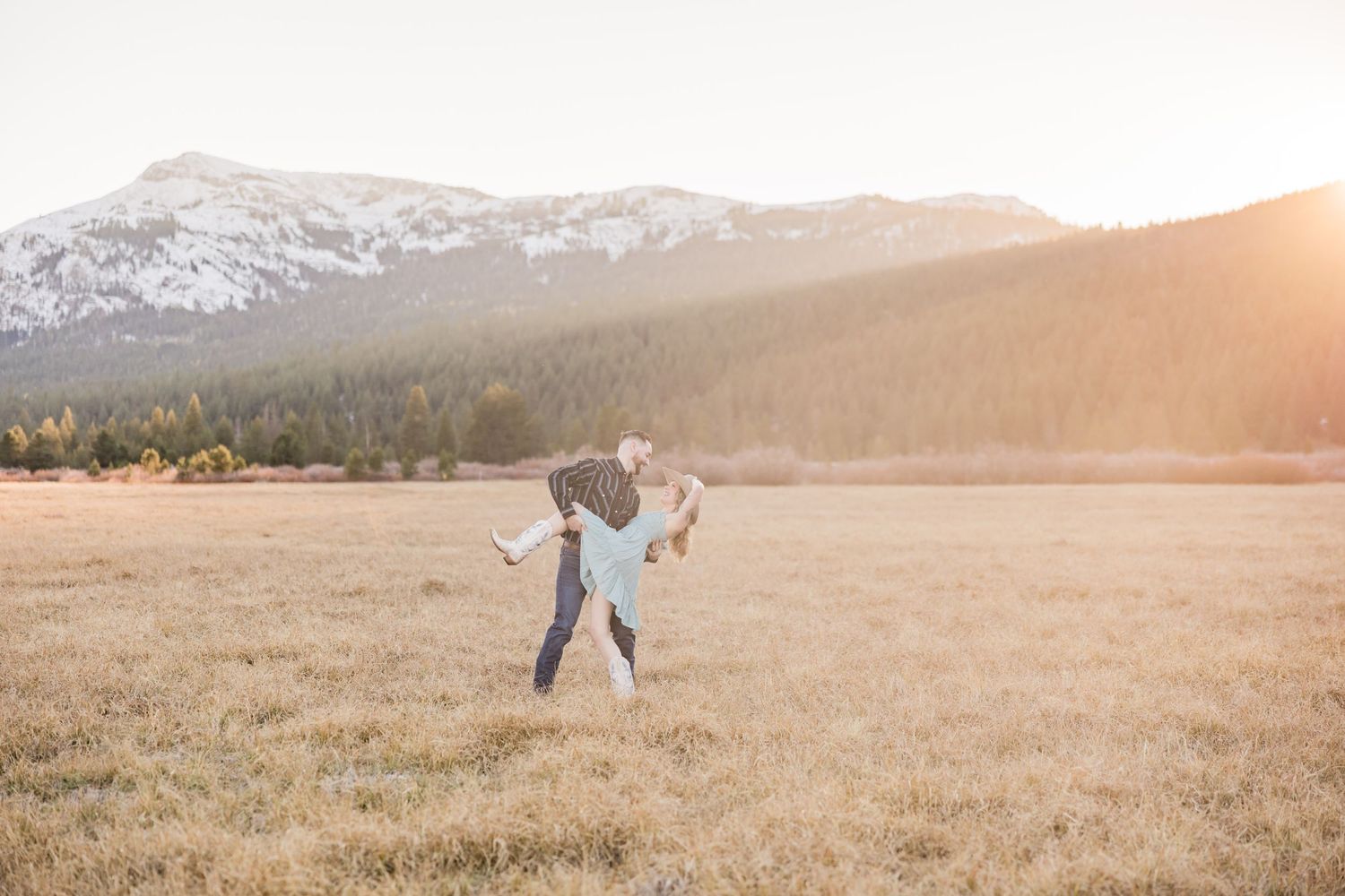 Couple holding hands running through golden meadow at sunset with mountain forest backdrop.