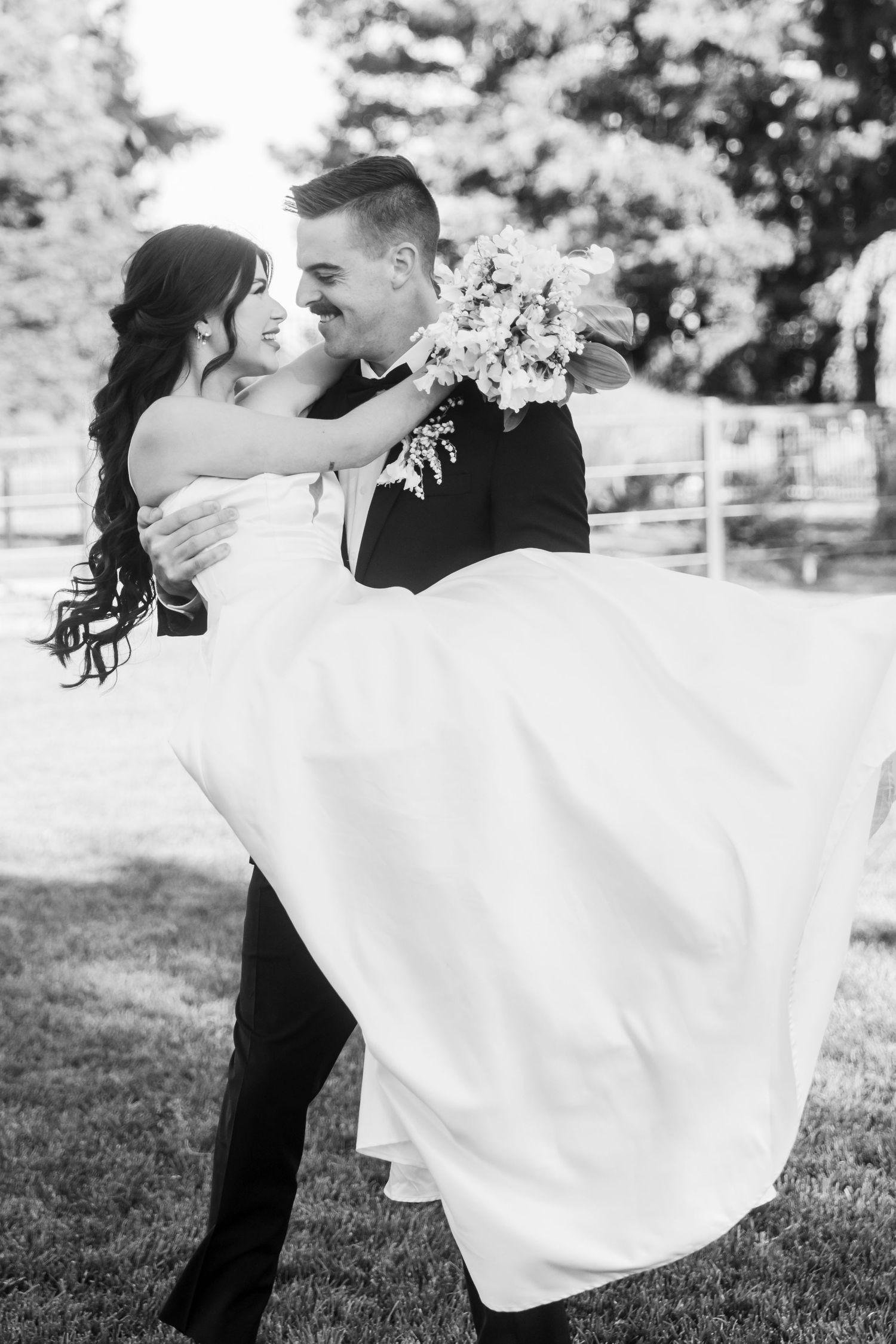 Black and white photo of a groom lifting his bride in a wedding dress outdoors, both smiling joyfully.