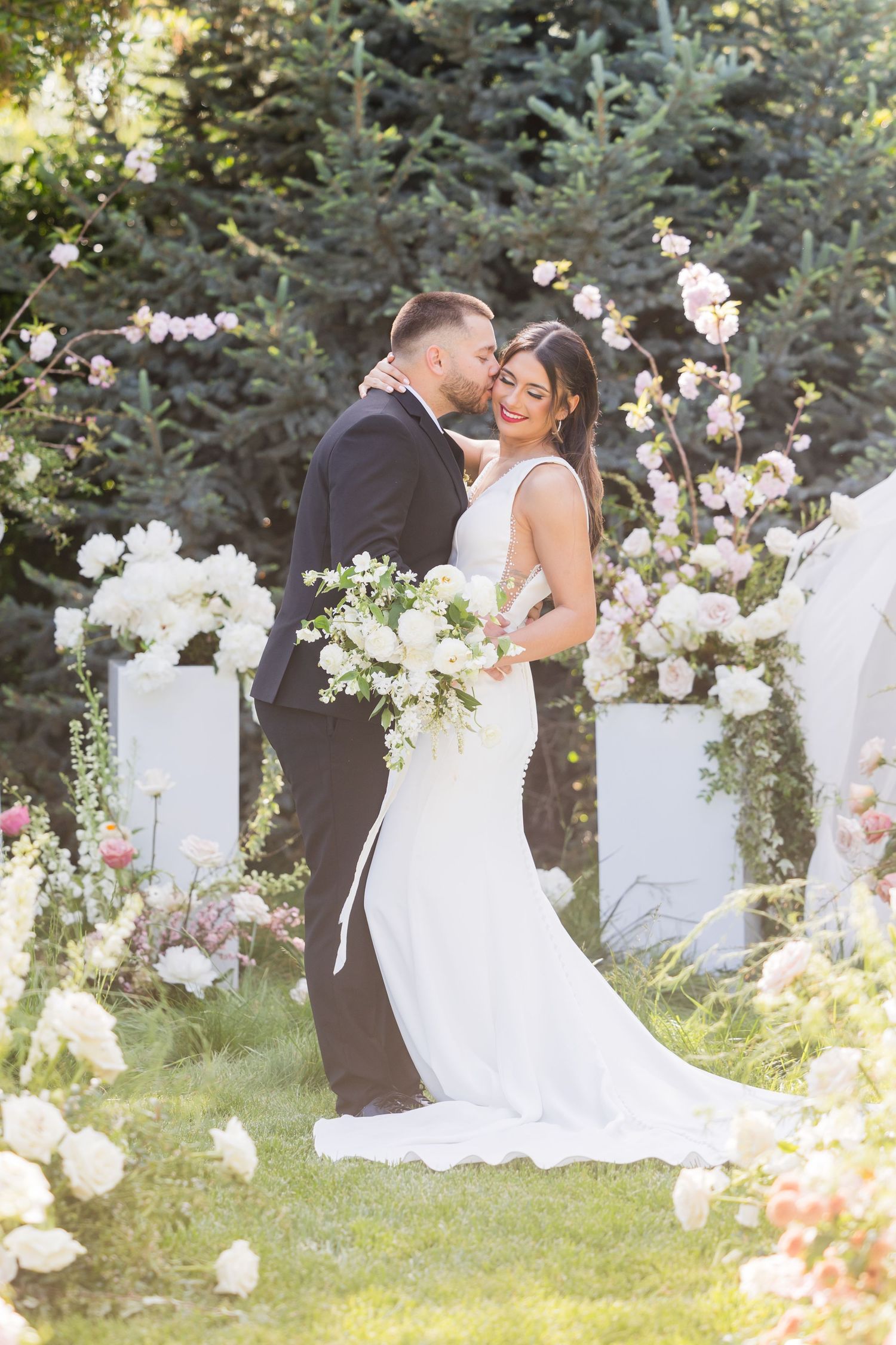 Bride and groom share a romantic kiss surrounded by lush white floral arrangements in a beautiful outdoor garden wedding ceremony.