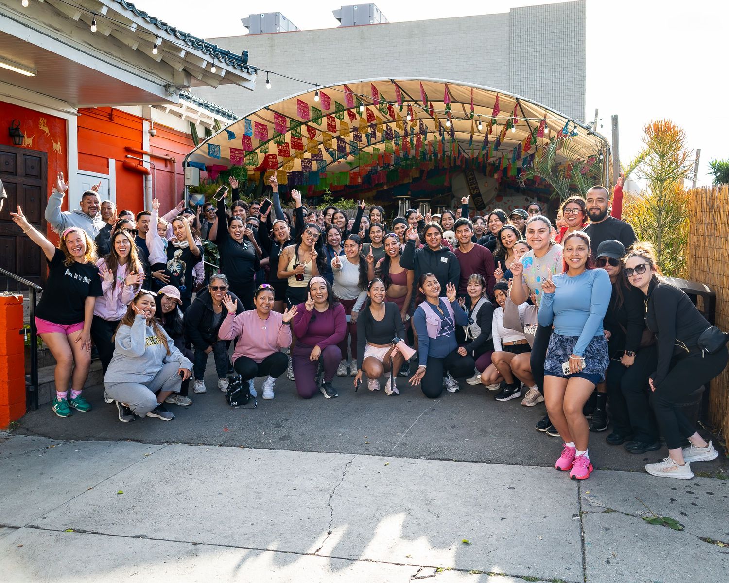 Large group posing and smiling in front of a colorful outdoor venue with streamers overhead.