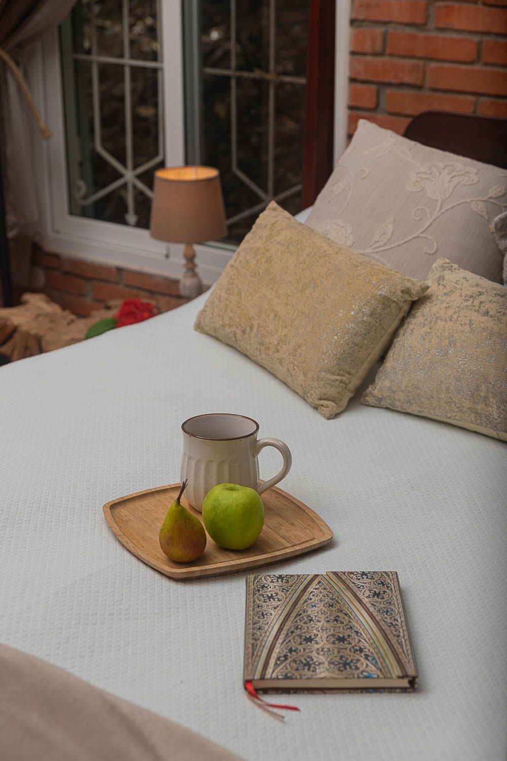 Cozy bedroom tray with ceramic mug, green apple, and pear beside ornate journal on white bed with decorative pillows.