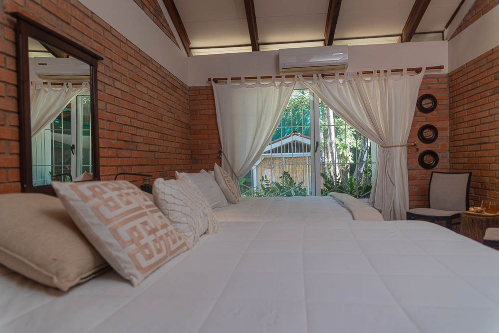 Cozy bedroom with brick walls, white bedding, decorative pillows, a window with curtains, and a wall-mounted AC unit.