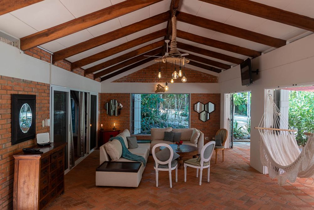 Open-air tropical living room with vaulted wood beam ceiling, hammock, chandelier, and lush garden views.