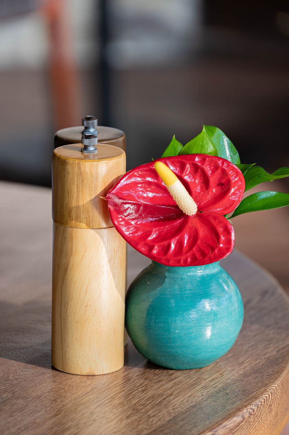 Red anthurium flower in teal vase beside wooden pepper grinder on table.