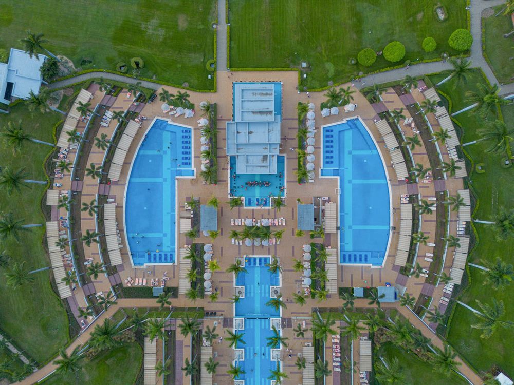 Aerial view of a luxury resort with symmetrical blue swimming pools, palm trees, and manicured green lawns.