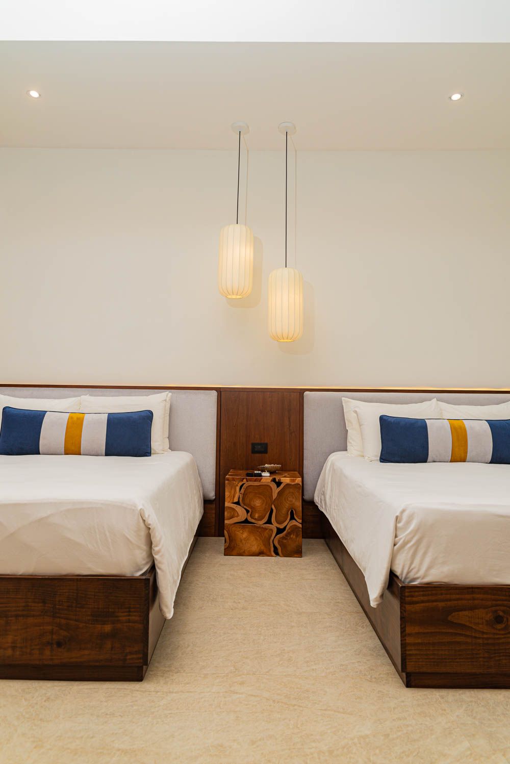 Two twin beds with white bedding and blue-gold pillows flank a wooden nightstand beneath pendant lights in a modern hotel room.