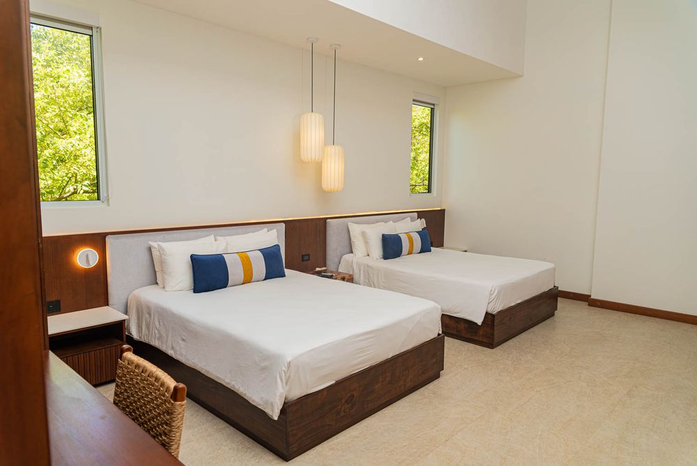 Modern hotel twin room with white bedding, colorful pillows, wooden headboard, pendant lights, and large windows.