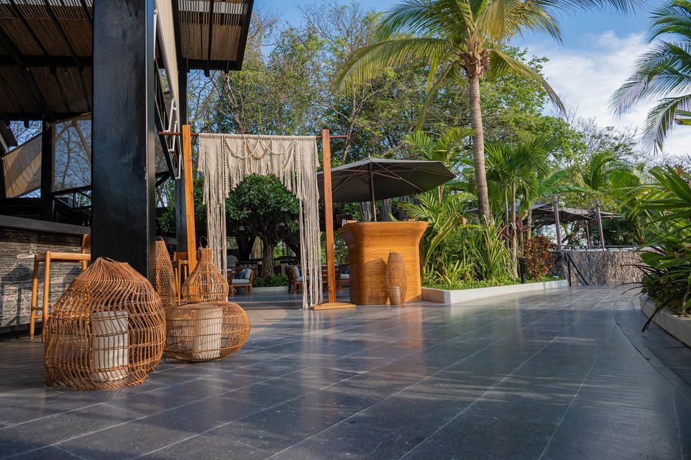 Open-air tropical venue with rattan decor, woven pendant chairs, and palm trees on a sleek dark tile patio.
