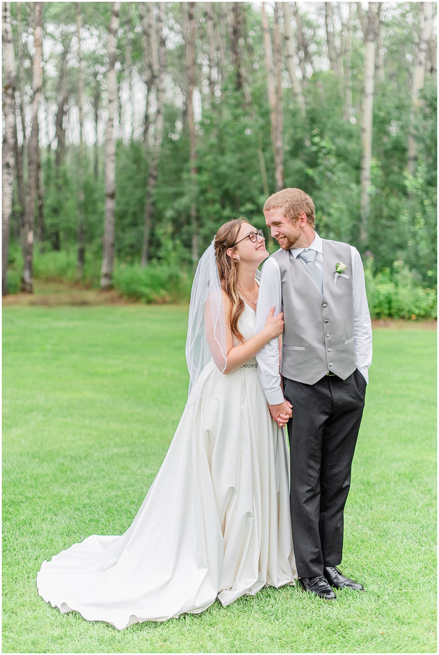 Bride and groom holding hands smiling at each other on green lawn with birch trees backdrop.
