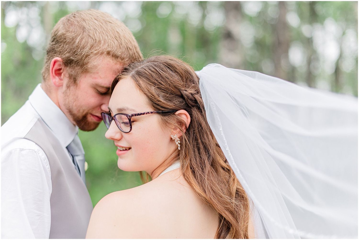 Groom nuzzles bride wearing glasses as her veil flows in the wind during outdoor wedding portraits.