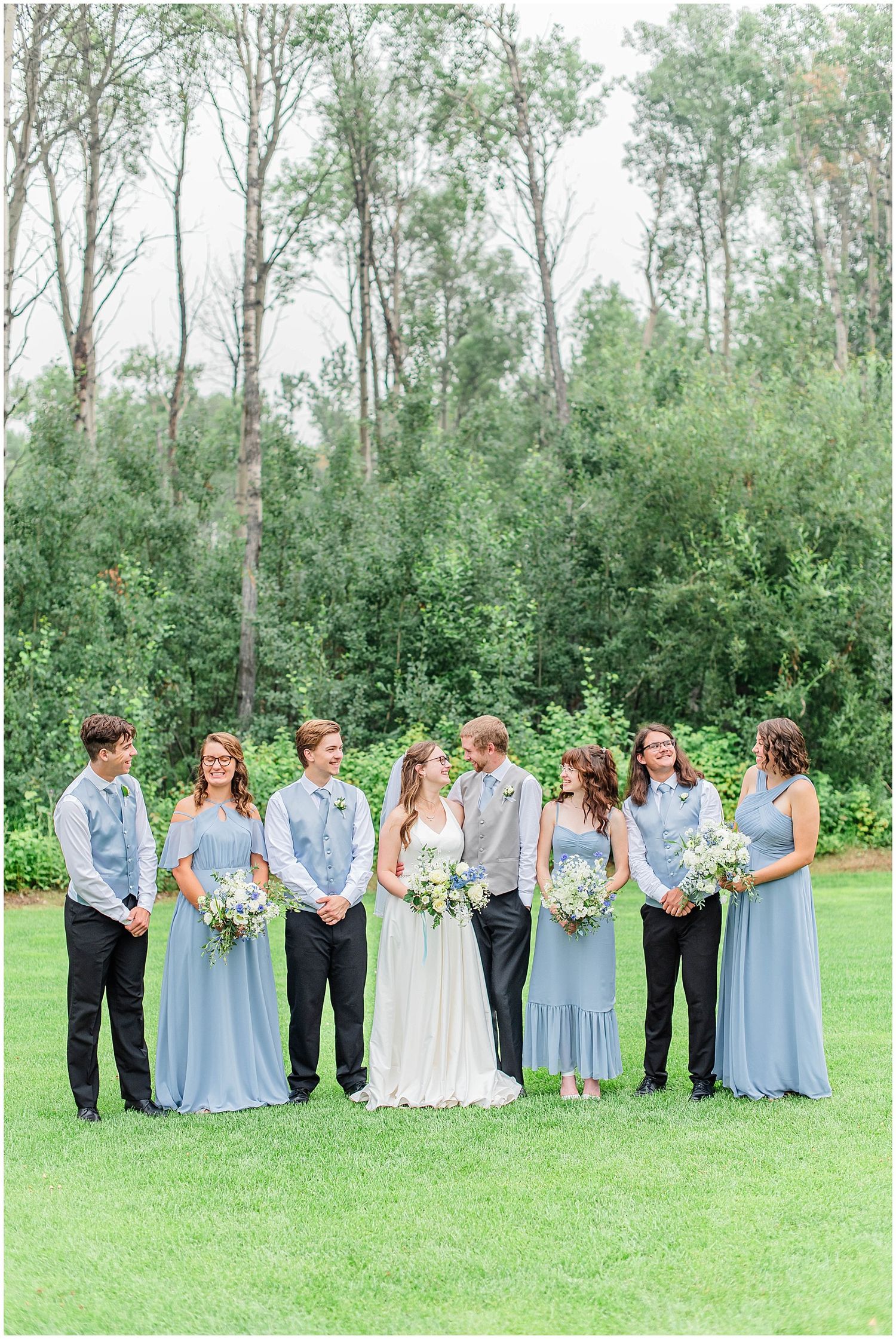 Wedding party in dusty blue dresses and black suits pose on green lawn with trees.
