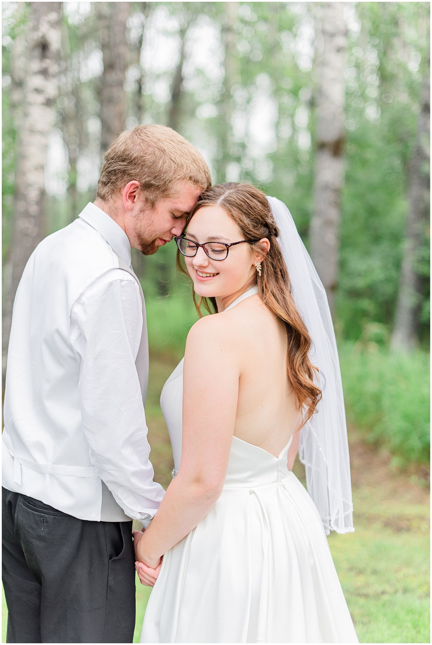 Smiling bride in backless gown and veil holding hands with groom outdoors.