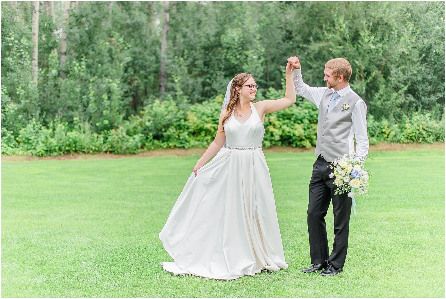 Groom spins his bride in an elegant white gown across a green lawn at their outdoor wedding.