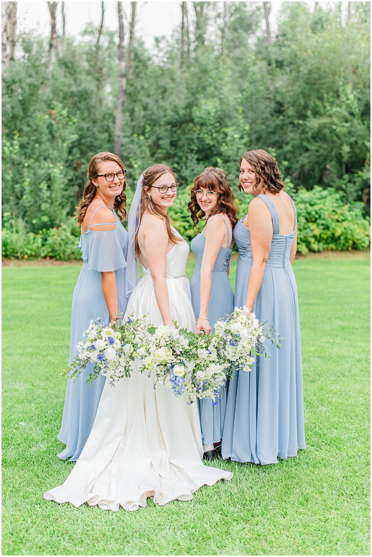 Bride poses with three bridesmaids in dusty blue gowns holding white floral bouquets outdoors.