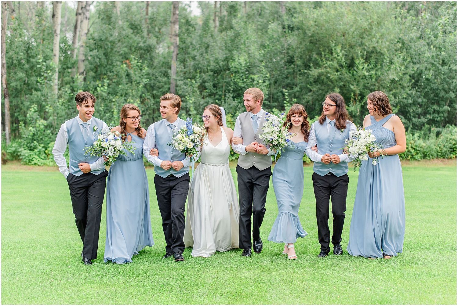 Wedding party walks together on green lawn with bridesmaids in dusty blue and groomsmen in gray.