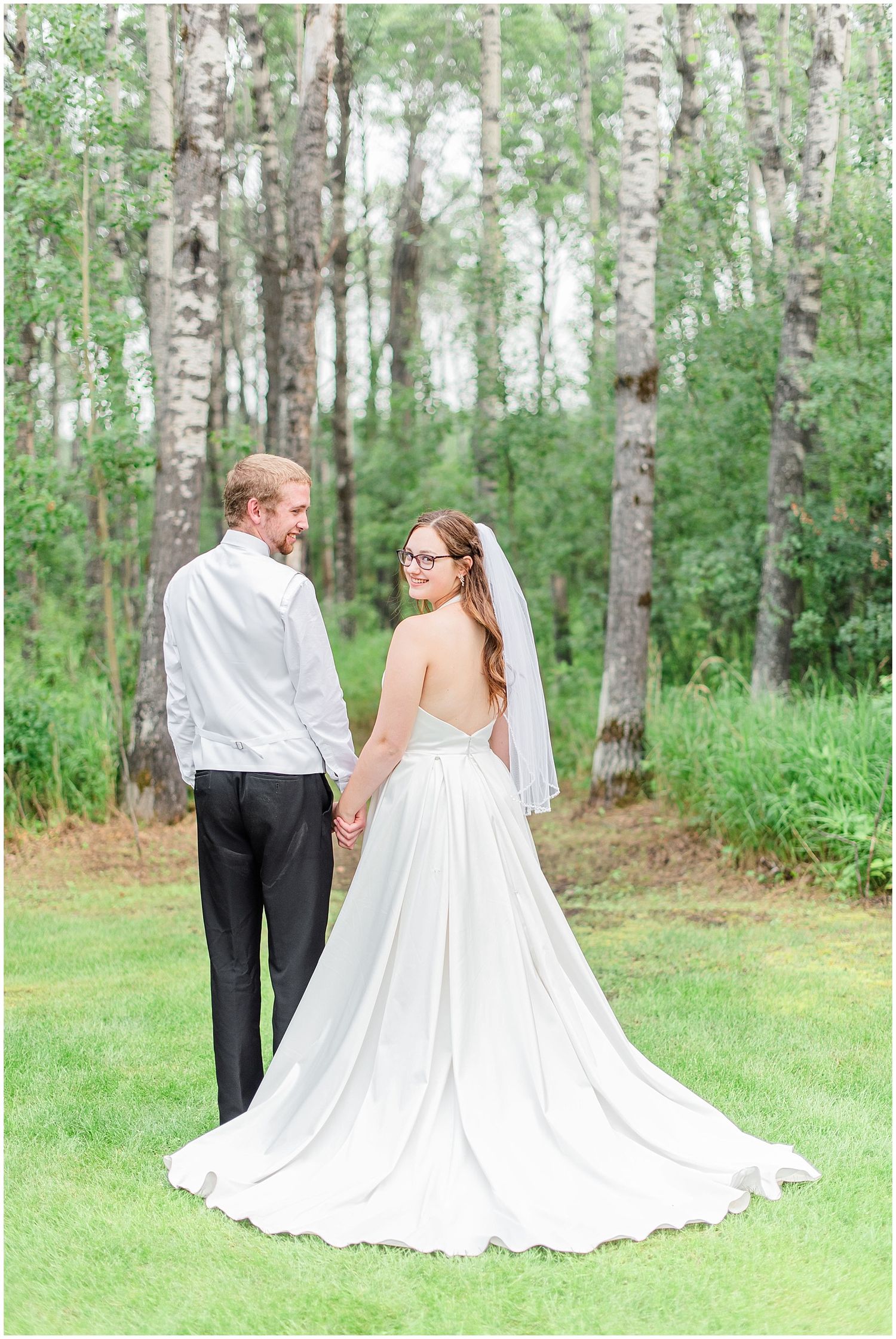 Bride and groom hold hands facing a lush birch tree forest, bride wearing elegant white gown with long train.