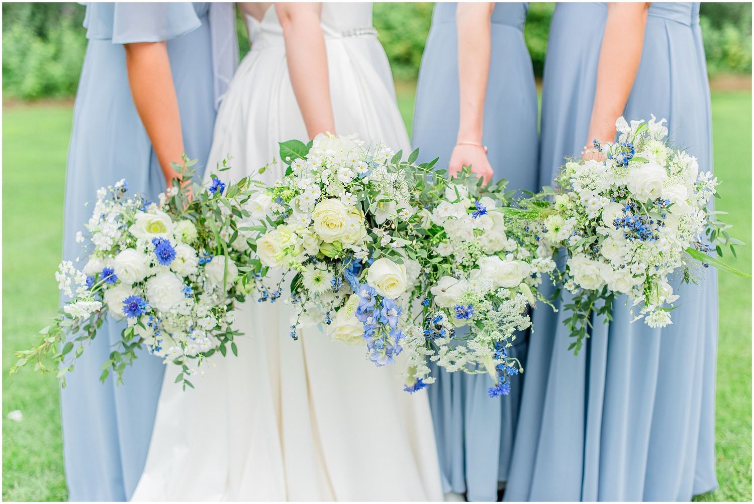 Bridesmaids in blue dresses holding lush white and blue floral bouquets with greenery at an outdoor wedding.
