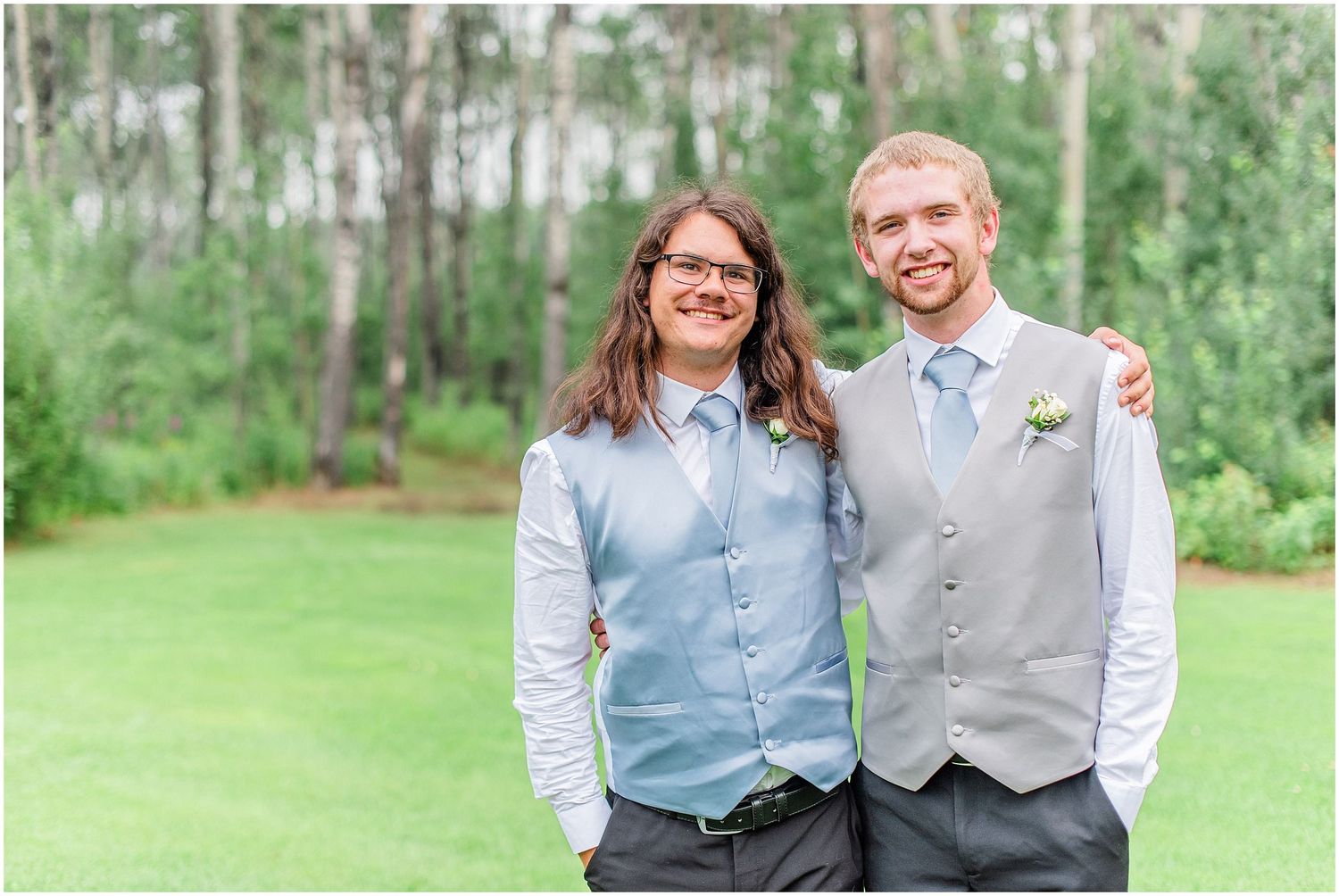 Two groomsmen smile together outdoors wearing light blue and gray vests at a wedding.