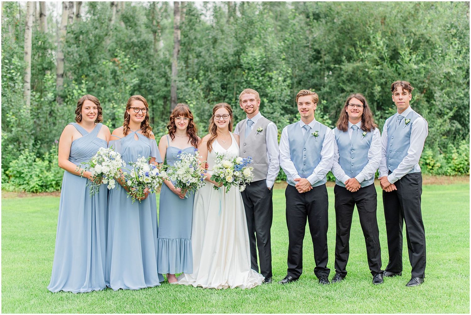Wedding party poses together on a green lawn, bridesmaids in dusty blue and groomsmen in gray.