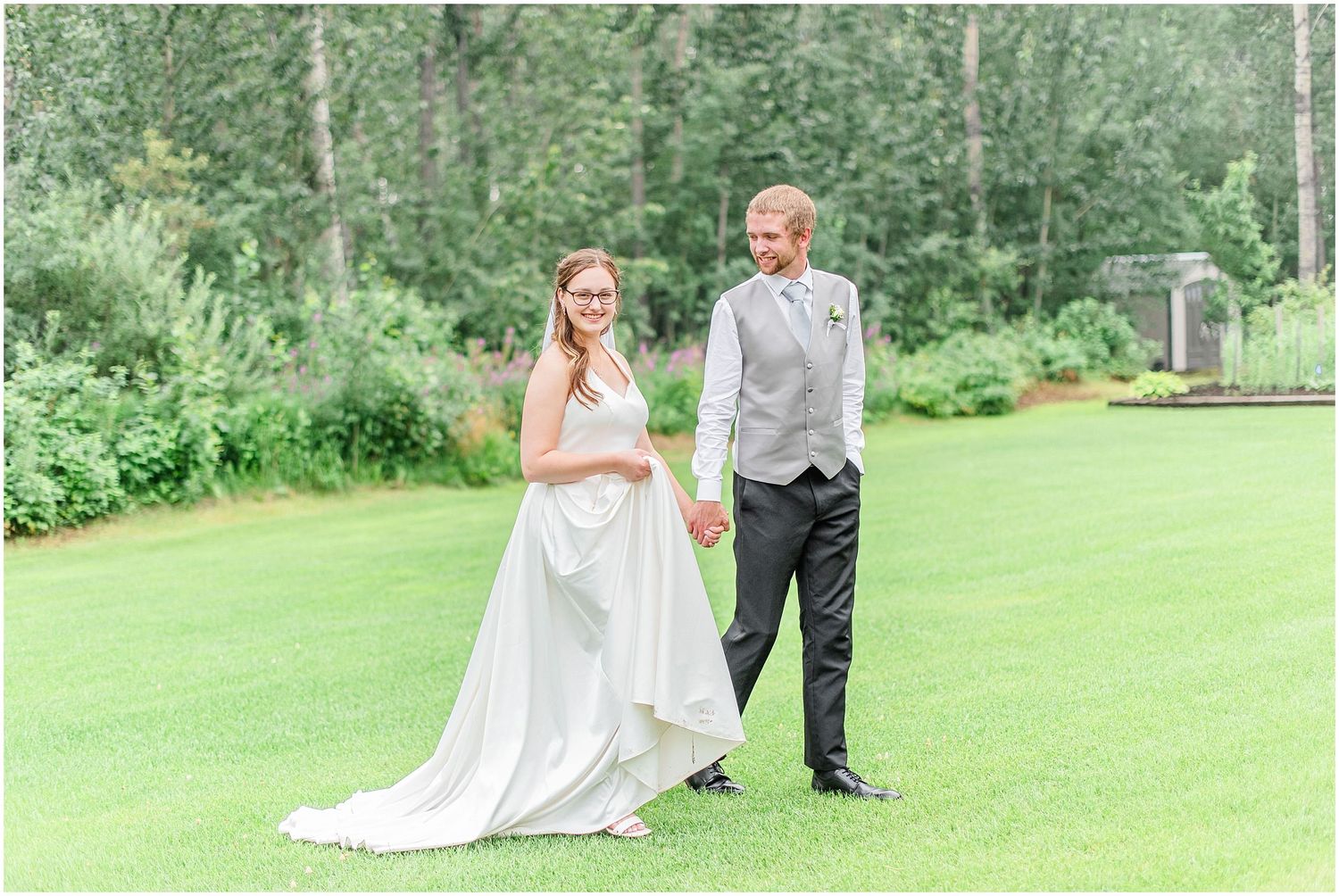Bride and groom holding hands on green lawn surrounded by lush trees at outdoor wedding ceremony.