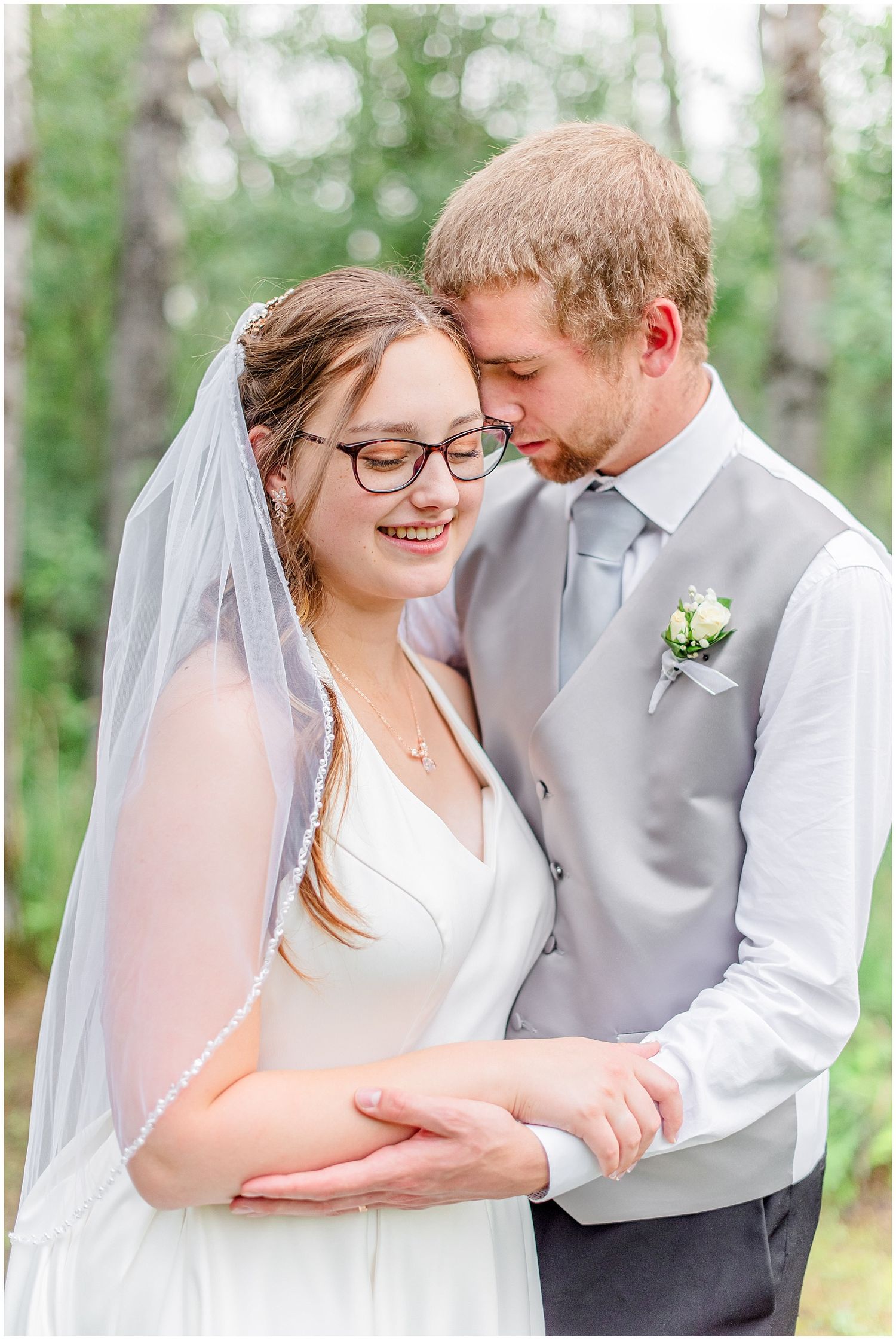 Bride wearing glasses and veil smiles as groom leans in close holding hands in wooded outdoor setting.