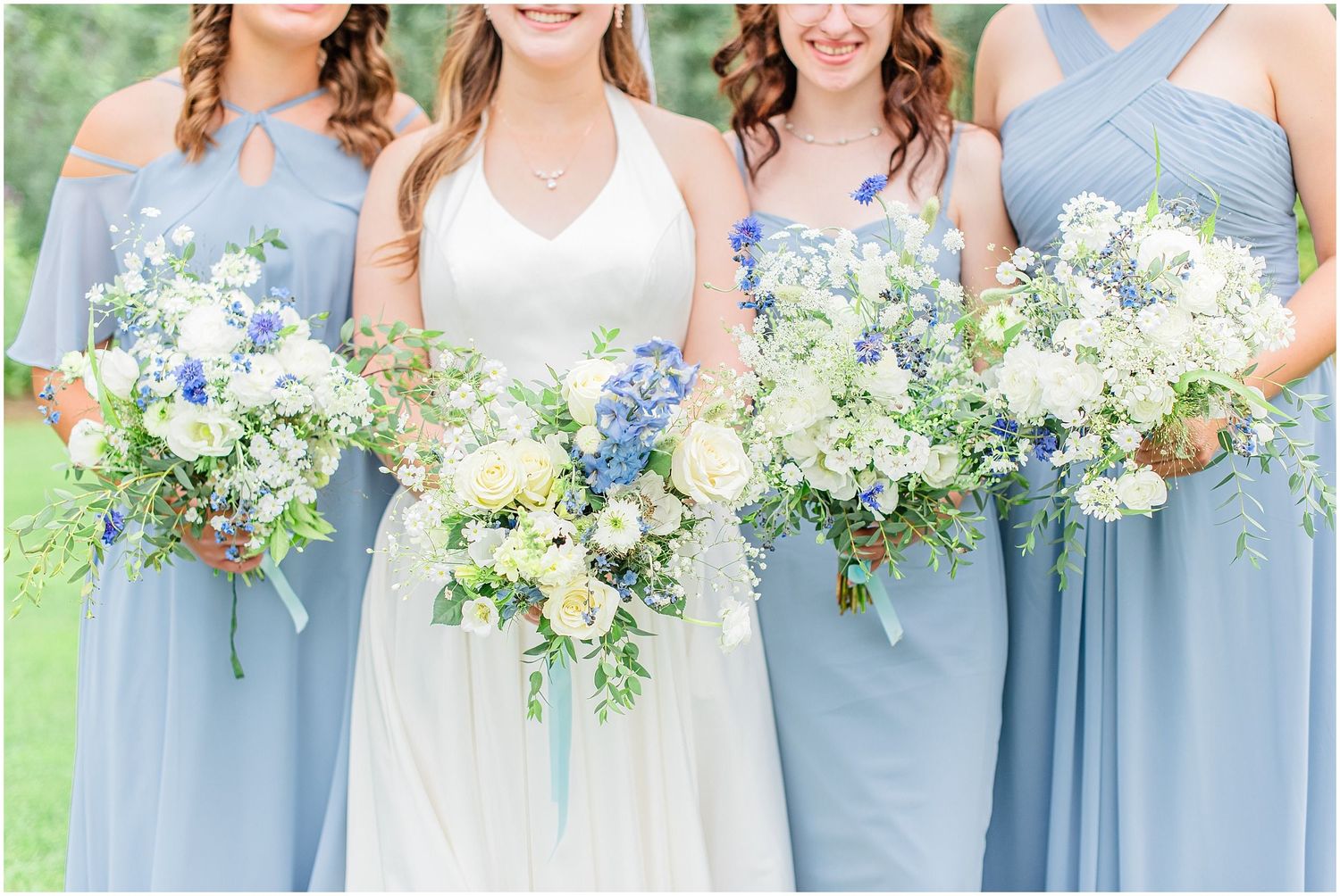 Bride and bridesmaids in dusty blue dresses holding white and blue floral bouquets outdoors.