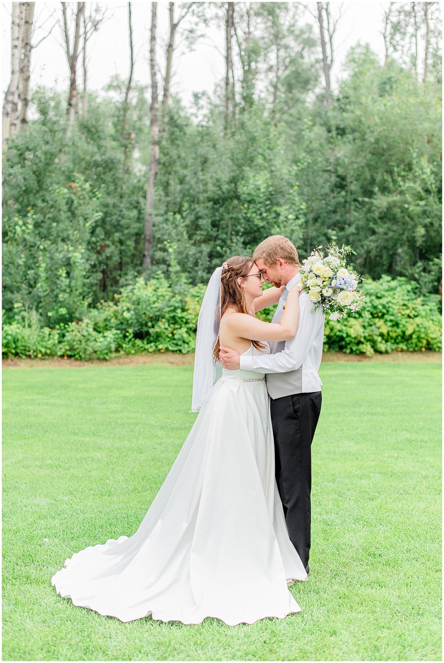 Bride and groom touching foreheads romantically on green lawn surrounded by lush garden and tall trees.