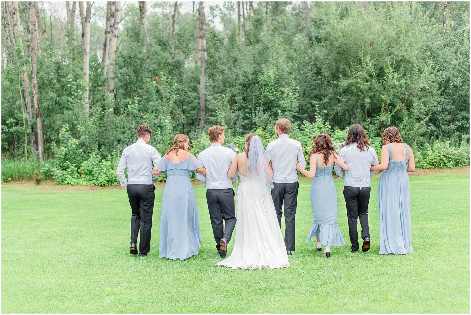 Wedding party walks away holding arms across a green lawn surrounded by lush trees.