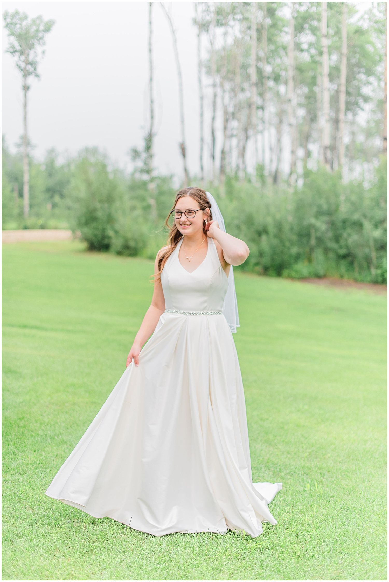 Bride in elegant white halter gown with veil smiling outdoors on green lawn surrounded by trees.