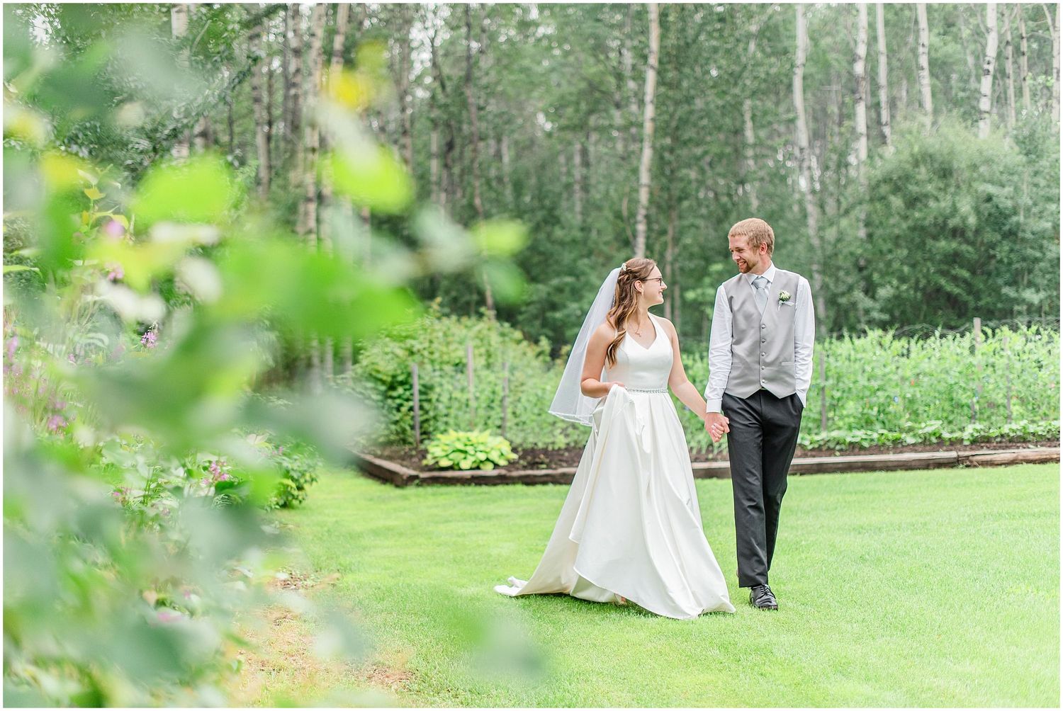 Bride and groom walking hand in hand through a lush green garden framed by vibrant foliage.