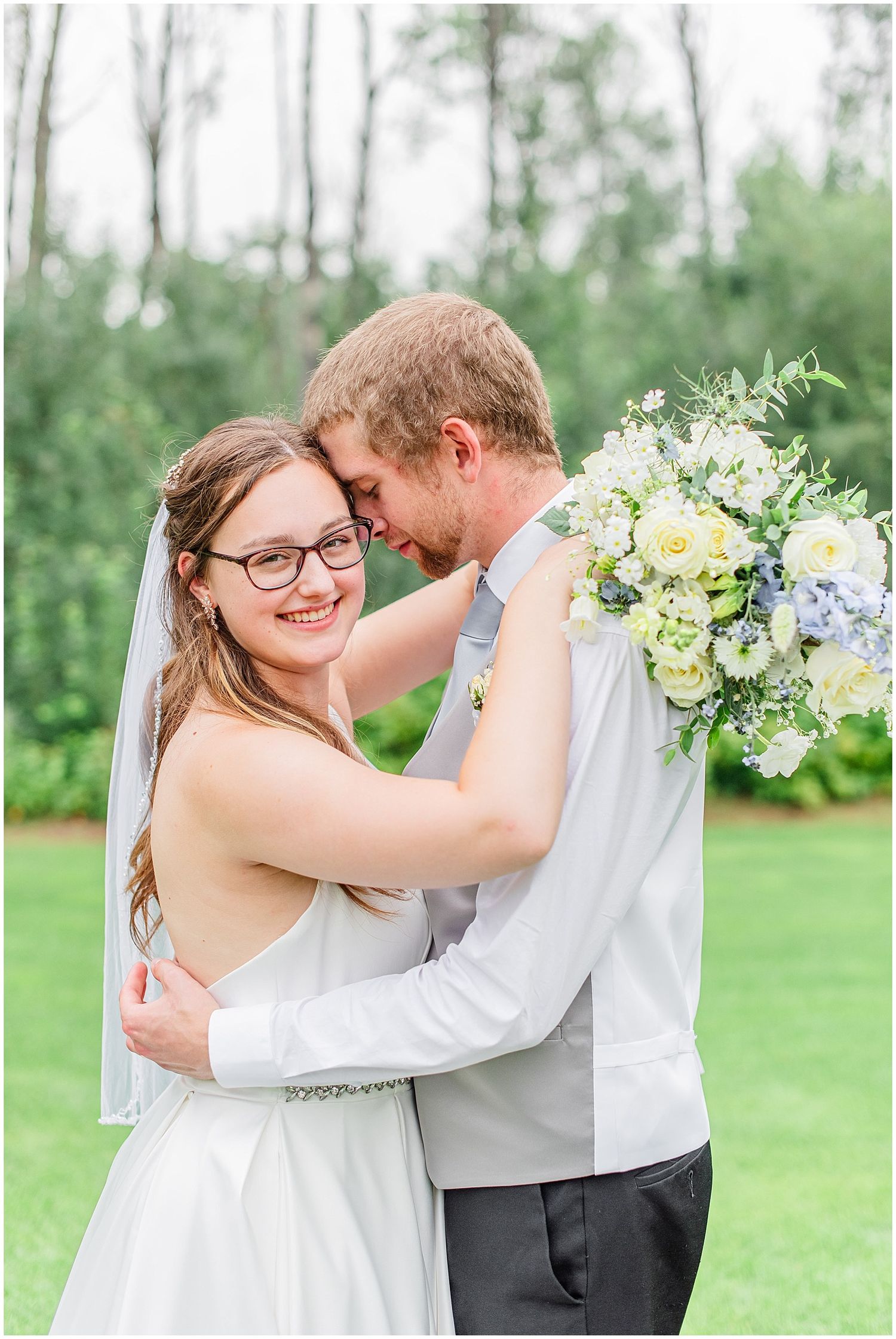 Bride and groom share a tender forehead touch while embracing outdoors, bride holding a blue and white floral bouquet.